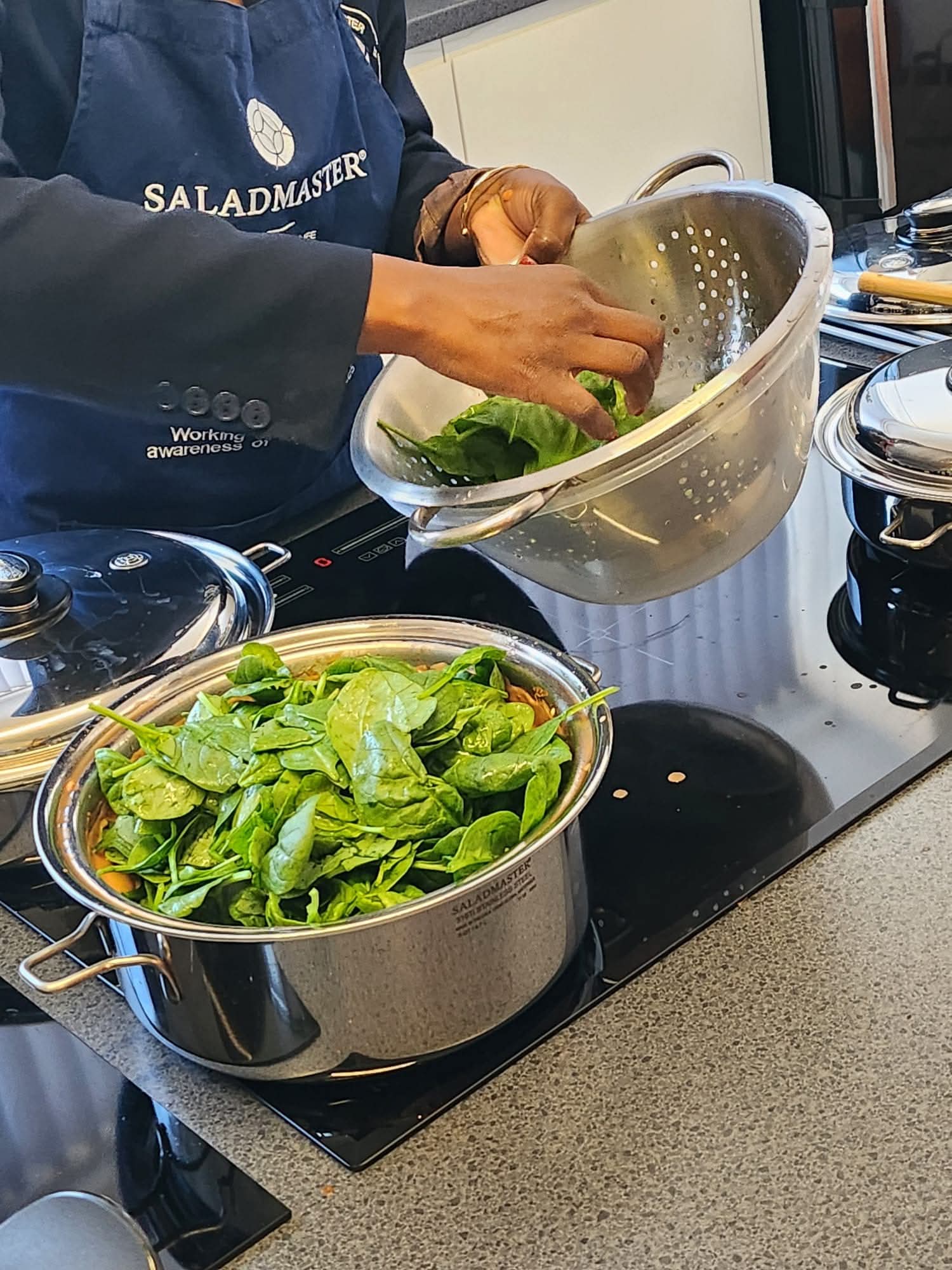 Fresh spinach being added to a Saladmaster stockpot with a colander during a live cooking demonstration